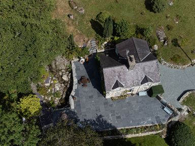 An aerial view of a stone house with slate roof and a large paved patio with outdoor furniture surrounded by grass and trees at Cae Lleci in Llanfachreth near Dolgellau