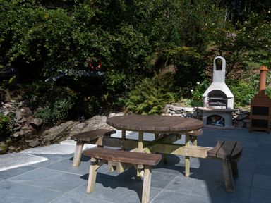 An outdoor patio with a round wooden picnic table and benches near a stone barbecue and a metal outdoor heater at Cae Lleci in Llanfachreth near Dolgellau