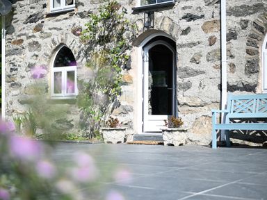 A stone building facade with an arched door and windows and a blue bench on a tiled patio at Cae Lleci in Llanfachreth near Dolgellau
