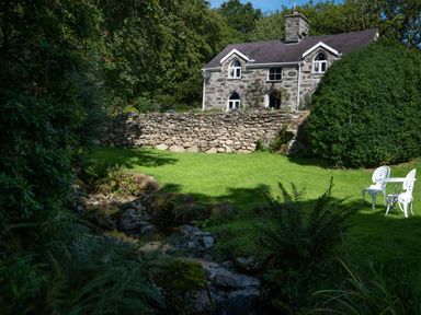 A stone house with a grassy garden and a white table with two chairs next to a small stream at Cae Lleci in Llanfachreth near Dolgellau