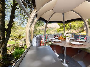 A round outdoor dining pod with cushions on bench seating a wooden table set with plates and a basket of fruit at Cae Lleci Llanfachreth near Dolgellau