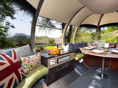 Interior of a covered outdoor dining area with curved benches cushions a table set with plates glasses a basket of fruit and scenic greenery outside at Cae Lleci in Llanfachreth near Dolgellau