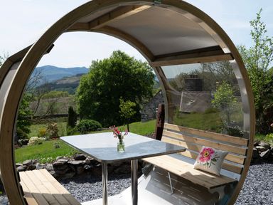 An outdoor seating area with a table and benches under a curved wooden shelter with a vase of flowers on the table at Cae Lleci in Llanfachreth near Dolgellau