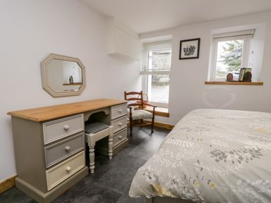 A bedroom with a wooden desk, chair, bed with floral bedding, two windows, and a framed picture on the wall at La'al Riddings in Lowick Green Riddings Farm Near Ulverston