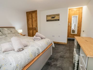 A bedroom with a bed, bedside table with lamp, wooden doors, a dresser with a stool, and a wall sign at La'al Riddings Lowick Green Riddings Farm Near Ulverston