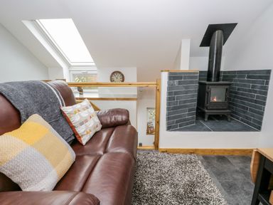 A sitting area with a brown leather sofa, two pillows, a gray blanket, a wood stove with black tiles, and a skylight window at La'al Riddings Lowick Green Riddings Farm Near Ulverston