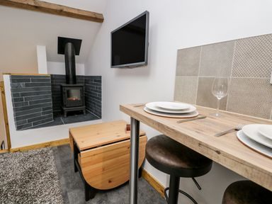 A dining area with a wooden table set for two with plates and a wine glass near a wall-mounted television and a wood-burning stove at La'al Riddings Lowick Green Riddings Farm Near Ulverston