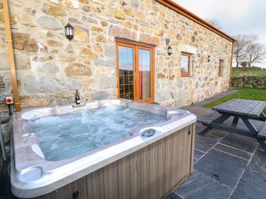 An outdoor hot tub with water bubbling next to a wooden picnic table on a stone patio outside a stone building with wooden framed doors and windows at Stabl in Pwllheli