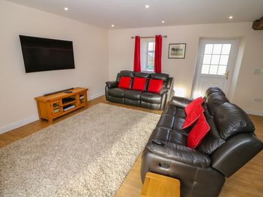 A living room with two black leather sofas with red cushions a wooden TV stand with a flat screen TV mounted on the wall and a beige rug at Stabl in Pwllheli