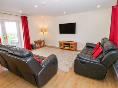 A living room with two black leather sofas red cushions a beige rug curtains a wooden tv stand with a mounted flat screen tv and a floor lamp at Stabl in Pwllheli
