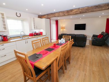 An open-plan kitchen and living area with a wooden dining table and chairs a kitchen counter with appliances and a black leather sofa set at Stabl in Pwllheli