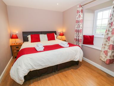 A bedroom with a bed with red pillows and a red runner two bedside tables with lamps and a window with floral curtains and a window seat with a red cushion at Stabl in Pwllheli
