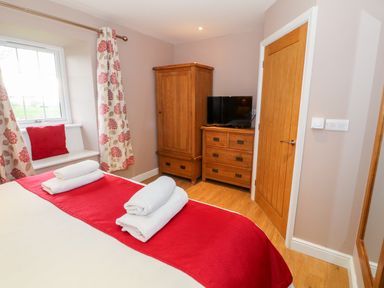 A bedroom with a bed with white and red bedding a window with floral curtains a wooden wardrobe and dresser with a television at Stabl in Pwllheli