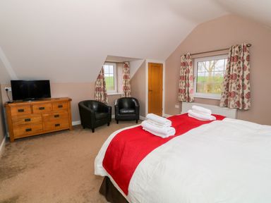 A bedroom with a bed with white linens and red runners two black armchairs a wooden dresser with a TV and windows with floral curtains at Stabl in Pwllheli