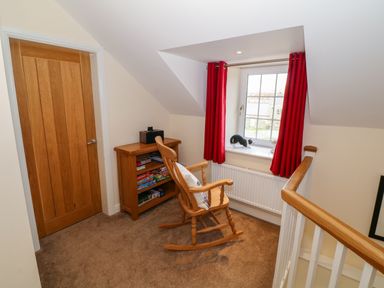 A corner room with a wooden rocking chair a small wooden bookshelf board games on shelves a window with red curtains and a small black and white cat on the windowsill at Stabl in Pwllheli