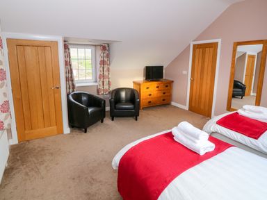 A bedroom with two beds with red blankets two black chairs a wooden dresser with a television a window with floral curtains and two wooden doors at Stabl in Pwllheli