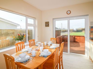 A dining area with a wooden table set with white dishes and mugs next to large windows and sliding glass door at The Trawler in Blackwater County Wexford