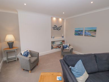A living room with grey armchairs, a blue sofa, a wooden side table with a lamp, wall art, and built-in shelves at Moorings in Kingswear Devon