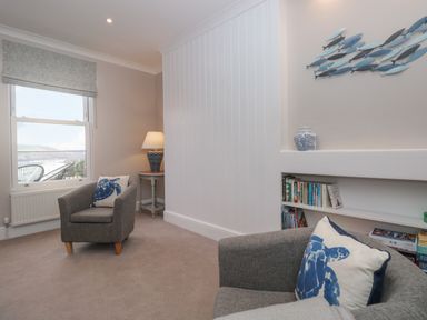 A sitting room with two grey armchairs with blue turtle cushions a side table with lamp and bookshelves at Moorings in Kingswear Devon