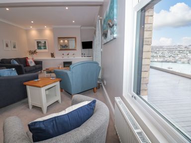 A living room with multiple chairs and sofas next to a large window overlooking a balcony and water at Moorings in Kingswear Devon