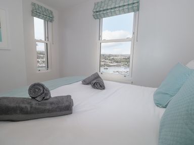 A bedroom with a bed with blue pillows and folded grey towels on it with two windows showing a view of water and buildings at Moorings in Kingswear Devon