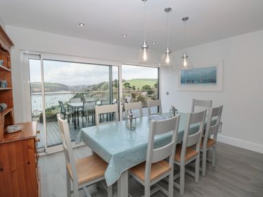 A dining room with a table covered with a tablecloth and eight chairs next to a sliding glass door with a view of an outdoor deck and a river at Moorings in Kingswear Devon