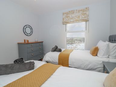 A bedroom with two single beds two gray rolled towels a gray chest of drawers and a window overlooking a harbor at Moorings in Kingswear Devon