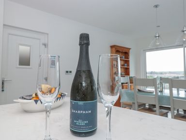 A kitchen counter with a bottle of sparkling wine and two empty glasses in a dining room with chairs and a window at Moorings in Kingswear Devon