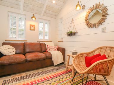 A living room with a brown leather sofa two pillows a wicker chair with two red pillows a wooden side table with a vase a round wooden mirror on the wall and hanging plants at Beach Hut in Trebarwith Strand