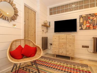 A room with a wicker chair and red cushions a patterned rug a wooden cabinet a wall-mounted TV a small shelf with a globe a wooden door and a round mirror at Beach Hut in Trebarwith Strand