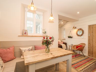 A dining area with a wooden table and built-in bench seating with pillows a wicker chair and mirror on the wall at Beach Hut in Trebarwith Strand