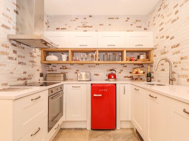 A kitchen with white cabinets a red mini fridge a red kettle toaster oven and open wooden shelves at Beach Hut in Trebarwith Strand