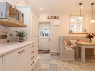 A kitchen corner with white cabinets a microwave a plant a wooden table with bench seating cushions windows and a door at Beach Hut in Trebarwith Strand