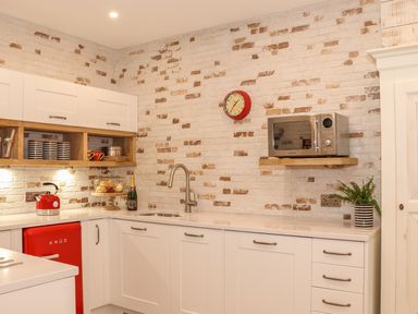 A kitchen with white cabinets a red mini fridge a red kettle a microwave on a wooden shelf a clock on a brick wall and a potted plant on the counter at Beach Hut in Trebarwith Strand