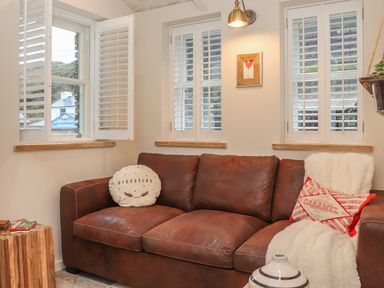 A brown leather sofa with cushions and a throw in a room with three windows with white shutters at Beach Hut in Trebarwith Strand