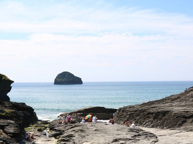 A rocky coastal area with people sitting on rocks near the sea and a small island in the distance at Beach Hut in Trebarwith Strand