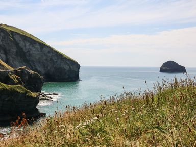 A coastal landscape with cliffs grassy foreground and a rock in the sea at Beach Hut in Trebarwith Strand