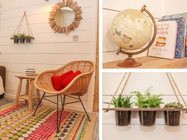 A corner of a room with a wicker chair with red cushions a wooden stool with a vase a hanging planter with three potted plants a round sunburst mirror and a globe with books on a shelf at Beach Hut in Trebarwith Strand