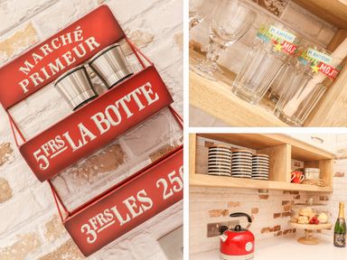 A kitchen wall with red metal shelves holding containers a wooden shelf with glasses and black striped mugs a countertop with a red kettle cake stand with pastries and a bottle at Beach Hut in Trebarwith Strand
