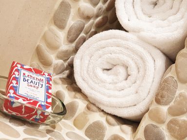Two rolled white towels and a bar of bath soap in a clear dish on a pebble stone countertop at Beach Hut in Trebarwith Strand