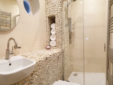 A bathroom with a sink a round mirror a pebble tiled wall with rolled towels and a glass shower enclosure at Beach Hut in Trebarwith Strand