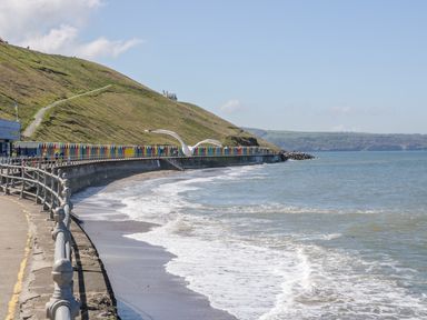 A coastal promenade with a railing along the sea and waves hitting the shore with green hills and colorful beach huts at West Cliff Apartment in Whitby