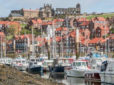 A harbor with boats moored at docks in front of red-roofed buildings and a large historic abbey on a hill at West Cliff Apartment in Whitby