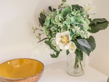 A glass vase with green and white flowers on a white surface next to a wooden bowl with a yellow interior at West Cliff Apartment Whitby