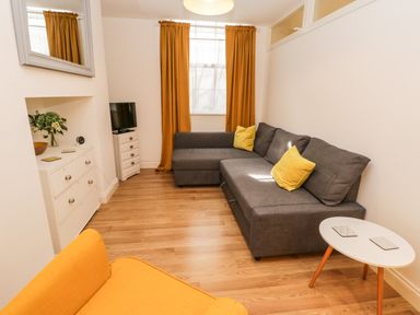 A living room with a gray sectional sofa with yellow cushions a yellow armchair a white round table wooden flooring and mustard curtains at West Cliff Apartment in Whitby