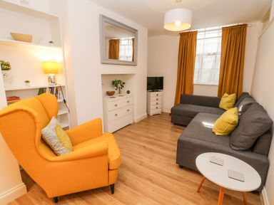 A living room with an orange armchair gray sofa a small white table and a TV on a stand at West Cliff Apartment in Whitby