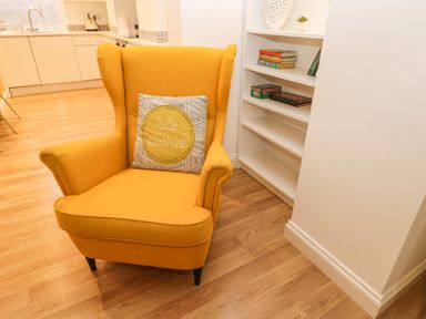 A yellow armchair with a patterned cushion next to a white bookshelf in a room with wooden flooring at West Cliff Apartment in Whitby