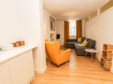 A living room with an orange armchair gray sofa yellow cushions a small round table and a shelf at West Cliff Apartment in Whitby