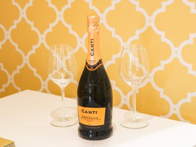 A bottle of Canti prosecco with two empty wine glasses on a white table with a yellow patterned wall in the background at West Cliff Apartment in Whitby