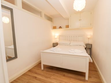 A bedroom with a white bed frame bedside tables with lamps a wall mirror and wooden flooring at West Cliff Apartment Whitby